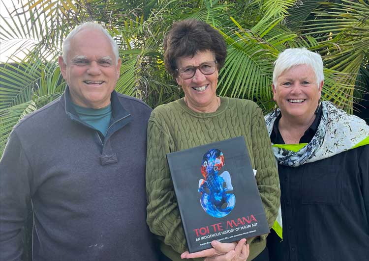 three older people smiling, middle one holding a book, palm trees in background
