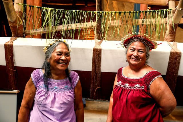 two elder Pacific women in colourful dresses with weaving gear, smiling