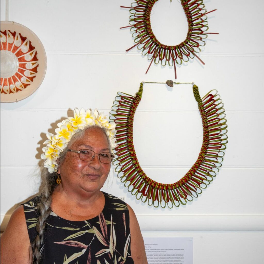 elder woman with yellow head dress and pacific necklaces on wall behind her
